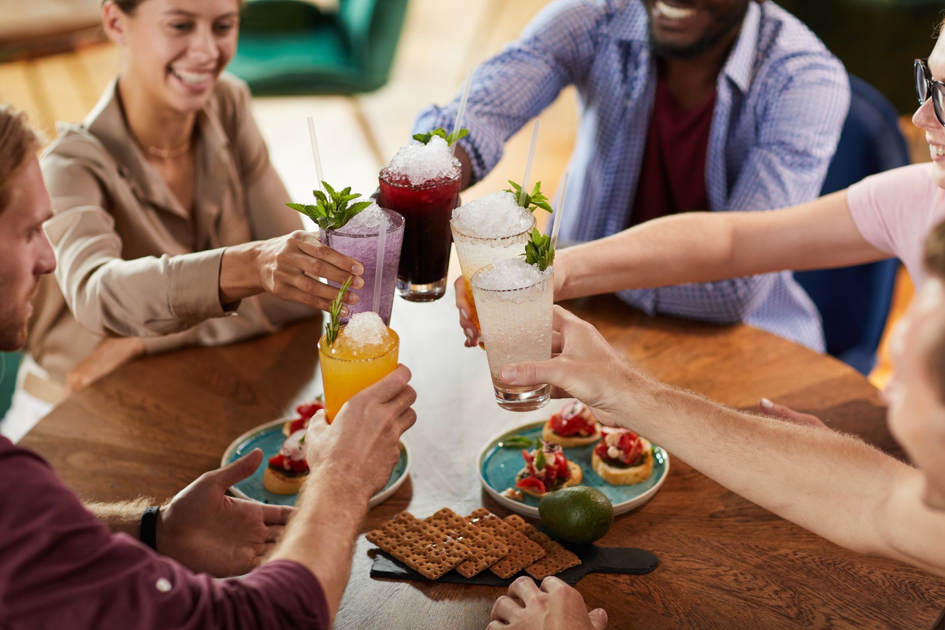 Group of people clinking colorful drinks over a table with appetizers and crackers.