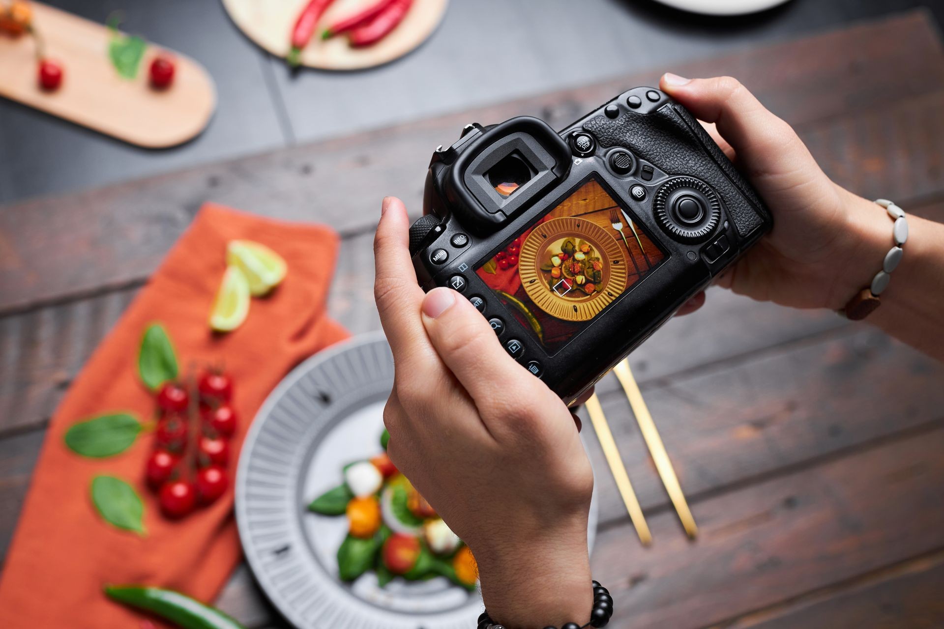 Person holding a camera displaying a food dish, surrounded by ingredients and utensils on a wooden table.